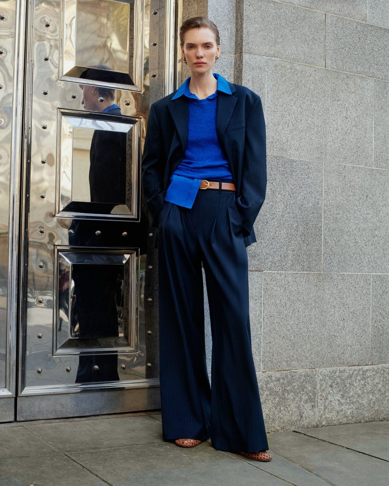 Model in navy suit and blue top standing against stone wall and metal door for ME+EM May Campaign