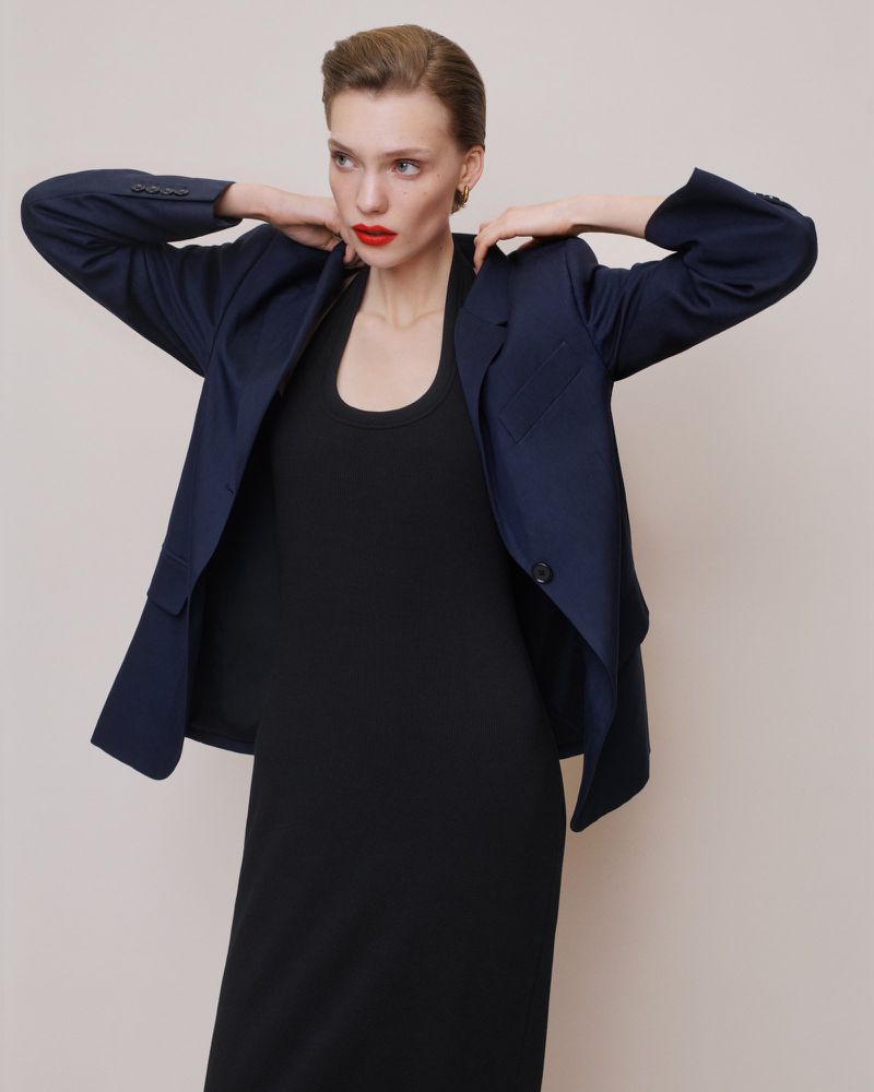 Model in a navy blazer over a black dress with red lipstick posing against a neutral grey studio backdrop.