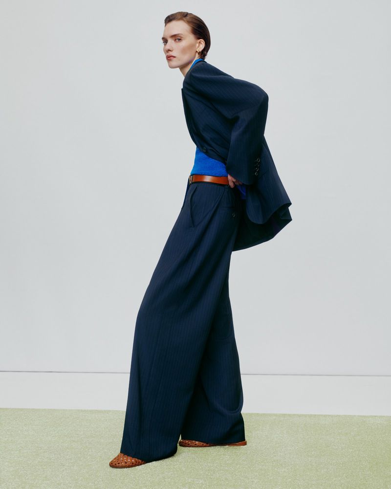 Model in navy blue oversized blazer and matching trousers with a bright blue top against a pale wall.