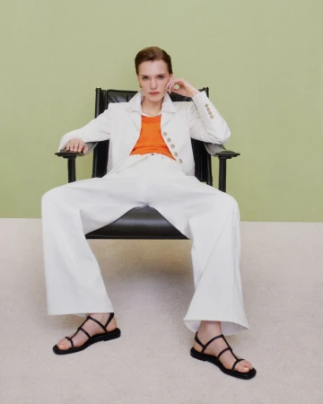Model wearing a white blazer and wide-leg trousers seated in a black leather armchair against a pale green wall.