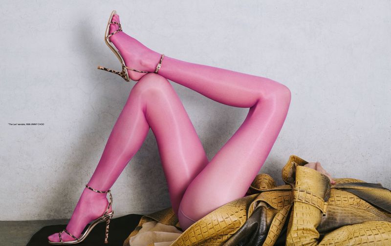 Woman's legs in pink stockings and strappy leopard-print high heels resting on a yellow crocodile-print jacket against a grey background.