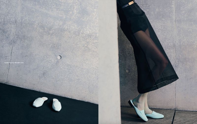 Fashion editorial featuring a woman leaning against a grey wall wearing black sheer pants and light blue-green loafers in a minimalist setting.