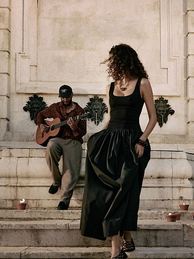 Woman in a black tank top and full dark skirt walking down stone steps past a man playing acoustic guitar