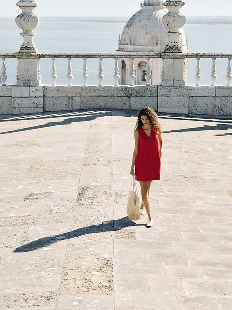 Woman in a red dress walks on a stone terrace, holding a straw bag, with a domed building and ocean in the background on a sunny day.