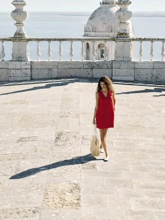 Woman in a red dress walks on a stone terrace, holding a straw bag, with a domed building and ocean in the background on a sunny day.