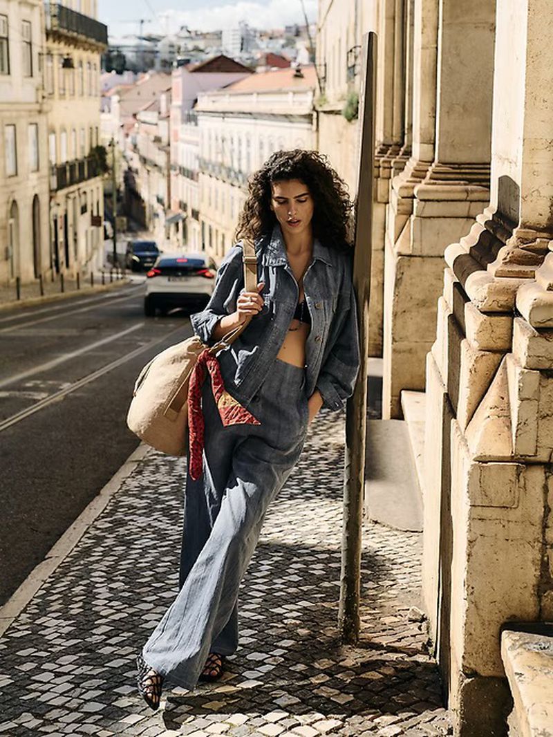 Model leaning against a stone building in a blue denim jacket and matching pants with a large canvas shoulder bag on a sunny sloped street