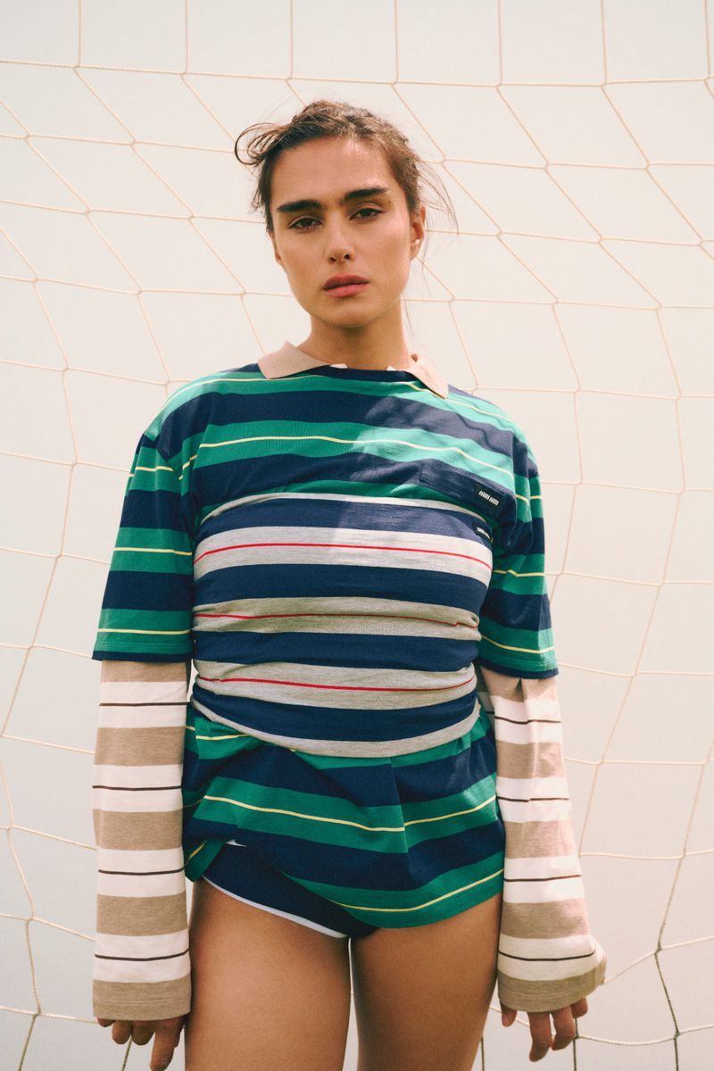 Brunette woman in striped shirt posing against a net. Fashion shot in natural light with athletic undertones and neutral backdrop.