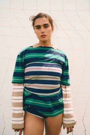 Brunette woman in striped shirt posing against a net. Fashion shot in natural light with athletic undertones and neutral backdrop.