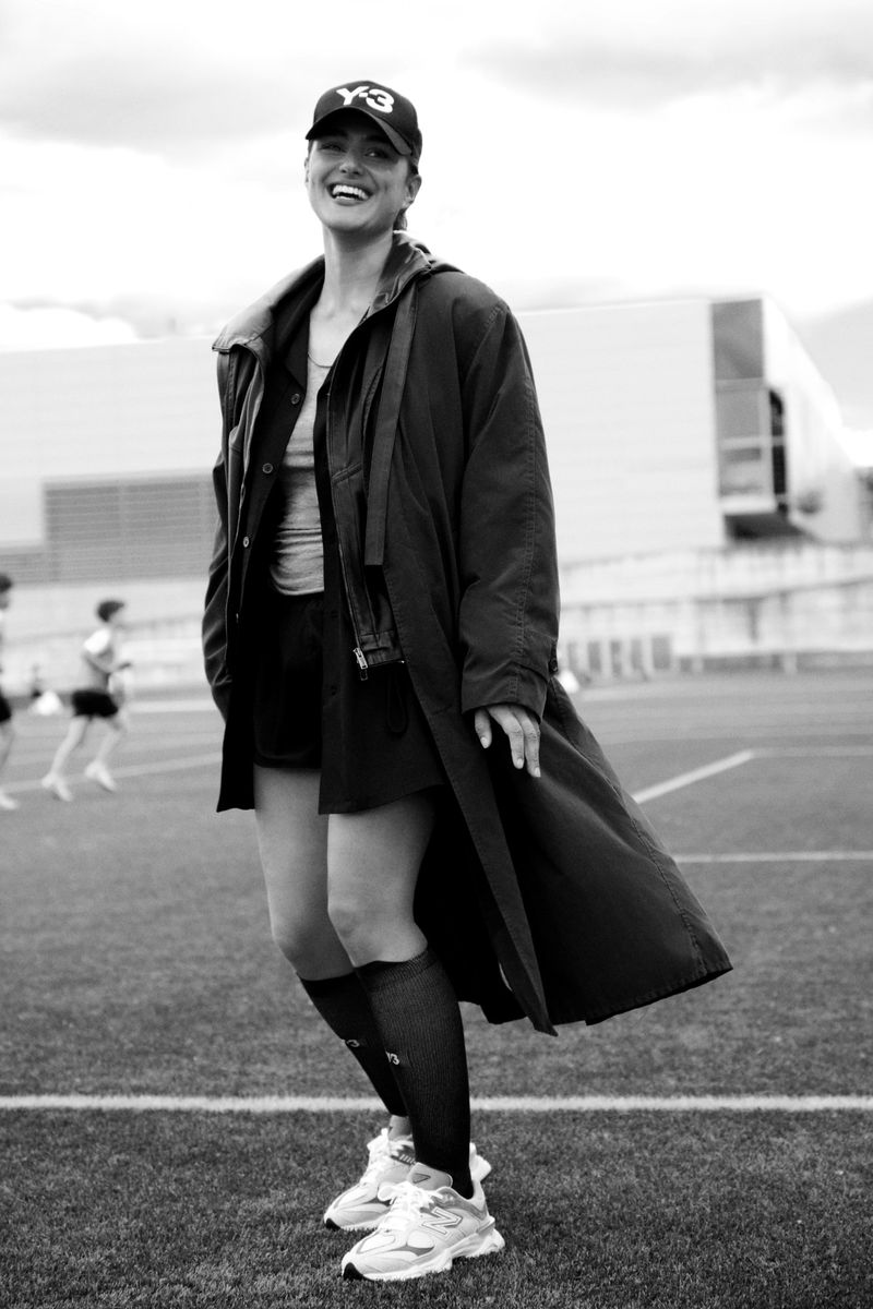Black and white portrait of a smiling woman in a baseball cap, jacket, and shorts on a grassy field with other people in the background.