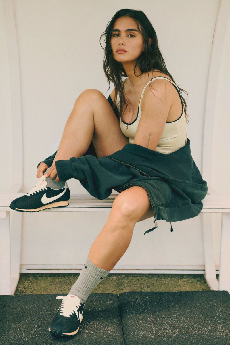 Woman sits on a bench outdoors wearing sportswear, gray socks, and tying the laces of black and white Nike sneakers.