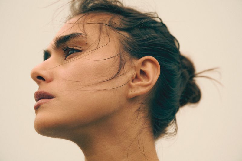 Close-up portrait of a young adult woman with dark hair in a bun, looking up with a neutral expression against a soft, neutral background.