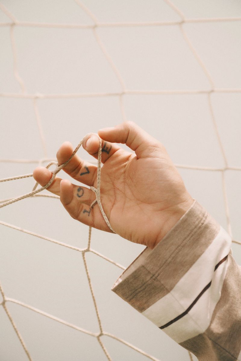 Close-up of an adult hand with visible tattoos holding a cream-colored sports net, with a neutral background and soft, diffused lighting.