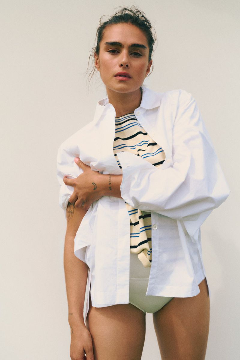 Woman in a white shirt and swimsuit bottom stands indoors against a neutral wall, with arms crossed, looking at the camera.