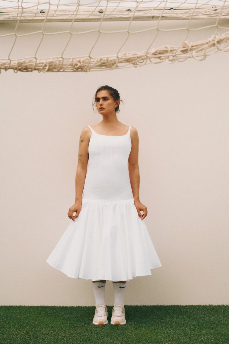 Woman in a white dress, athletic socks, and sneakers stands on artificial grass in front of a goal net and neutral wall.