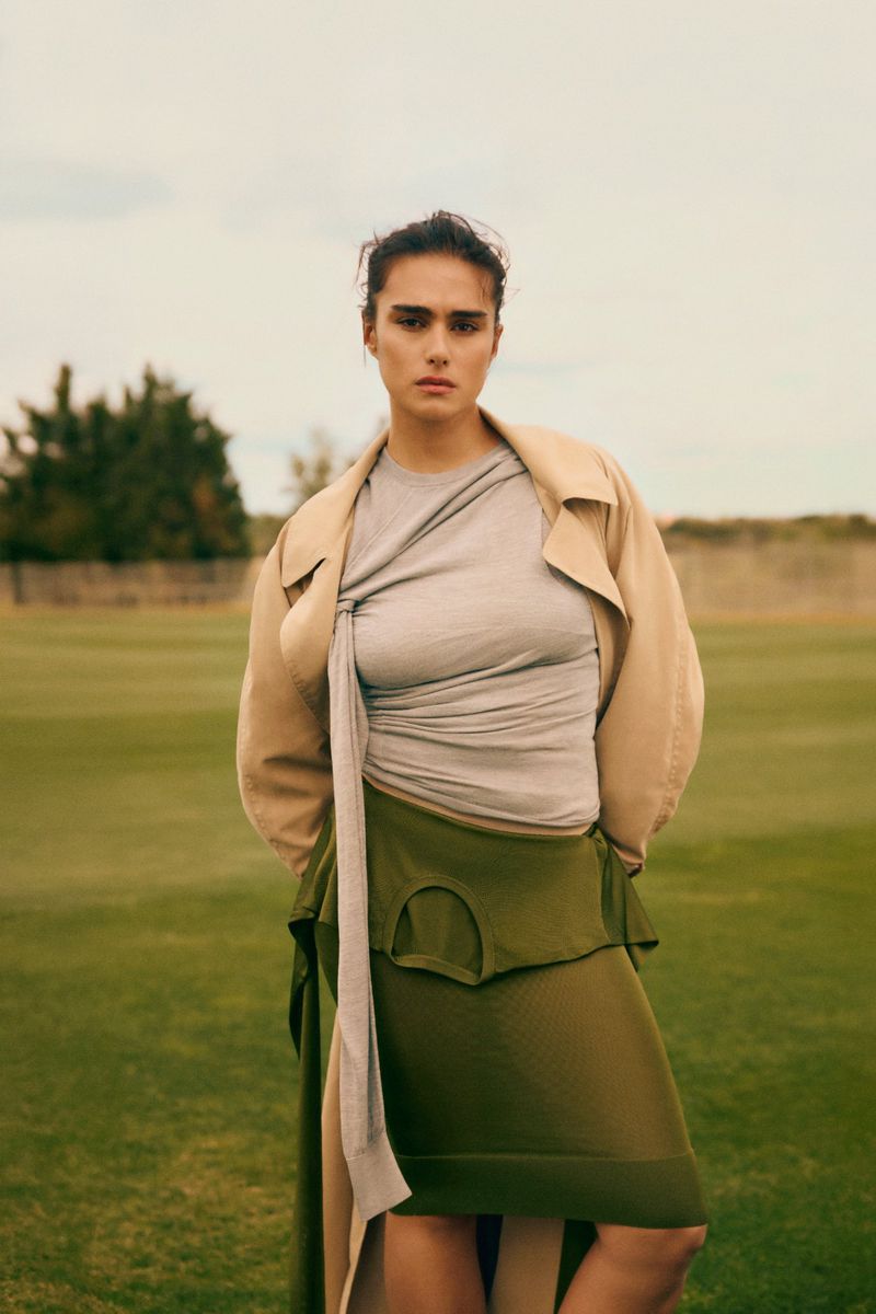 Model Jill Kortleve posing outdoors on a green lawn wearing a draped grey top, olive skirt, and beige coat for Vogue España.
