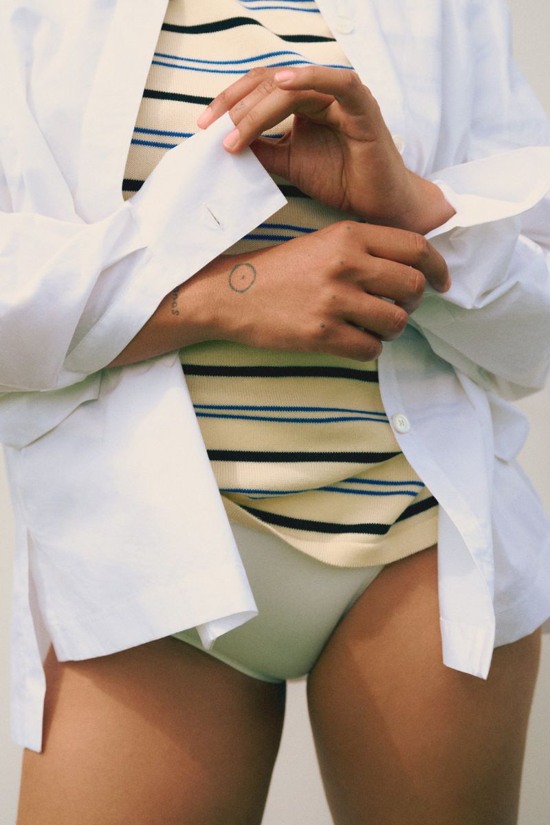 A woman models a striped top and white briefs, wearing a white shirt, indoors with soft lighting in a close-up fashion shot.
