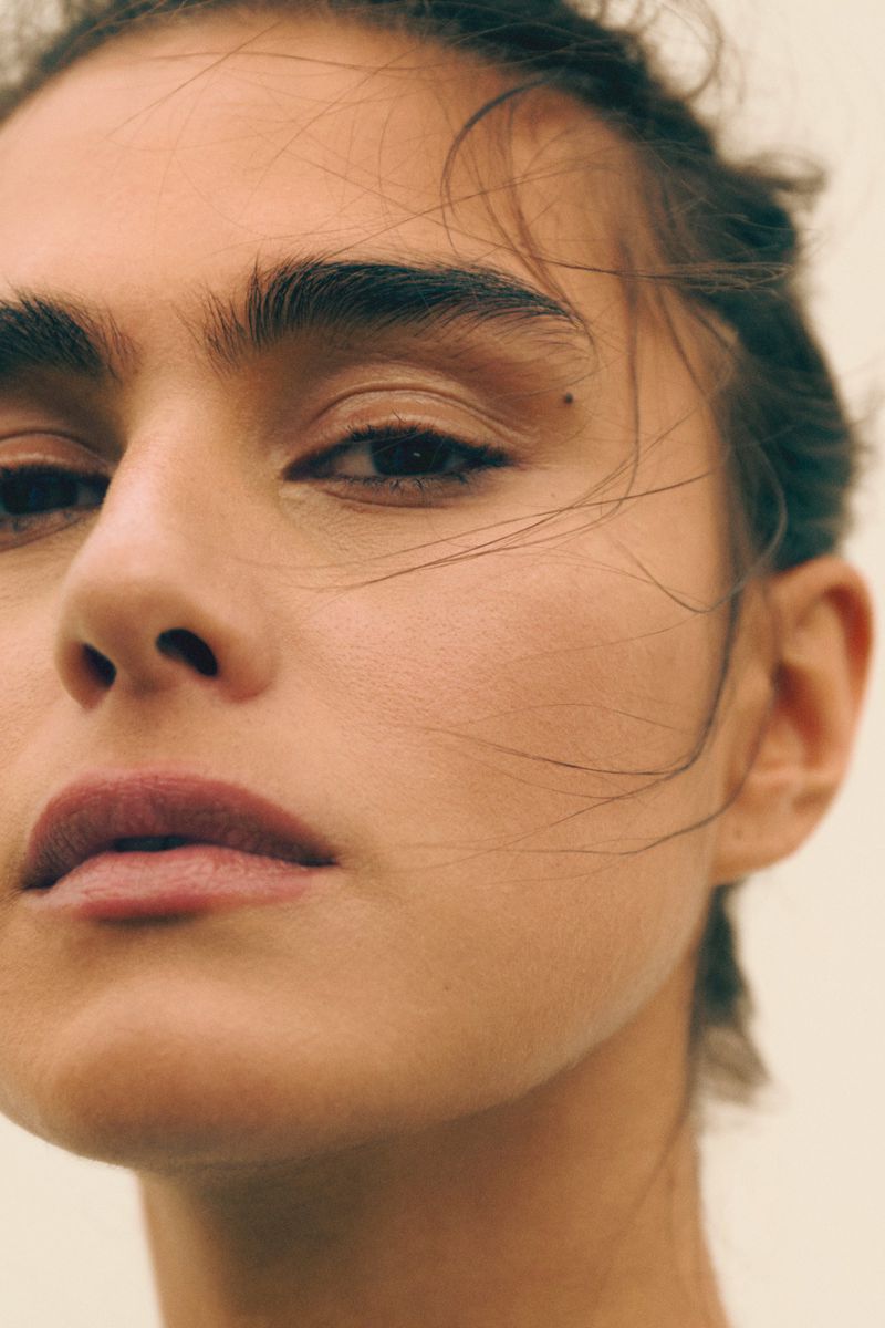 Close-up portrait of a woman's face featuring fair skin, dark eyes, defined eyebrows, and a neutral expression against a soft background.