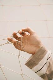 Close-up of a hand with finger tattoos spelling LOVE gripping a soccer net while wearing a beige and white striped jersey sleeve.