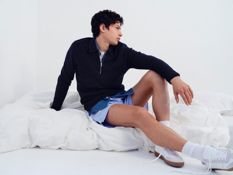 Model sitting on white bedding wearing a dark zip sweater, blue striped shorts, and white sneakers against a white background