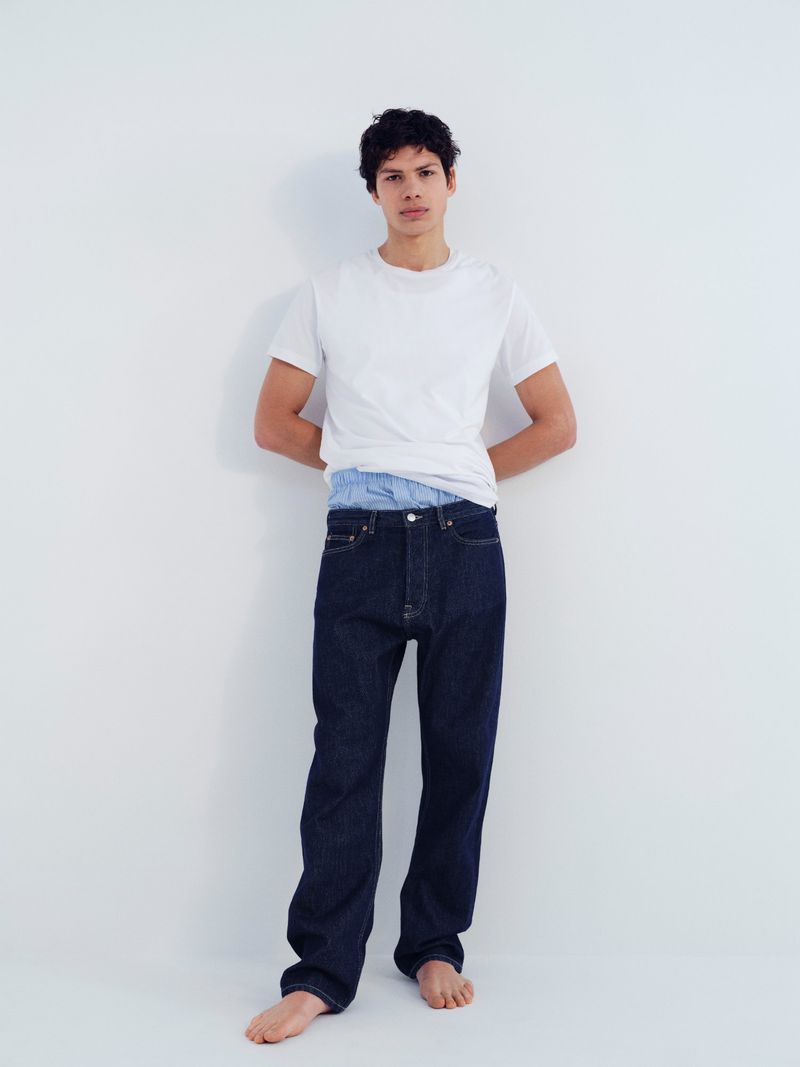 Full-length portrait of a male model wearing a white crewneck t-shirt and dark blue jeans against a plain white background.
