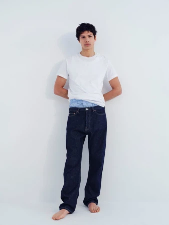 Full-length portrait of a male model wearing a white crewneck t-shirt and dark blue jeans against a plain white background.