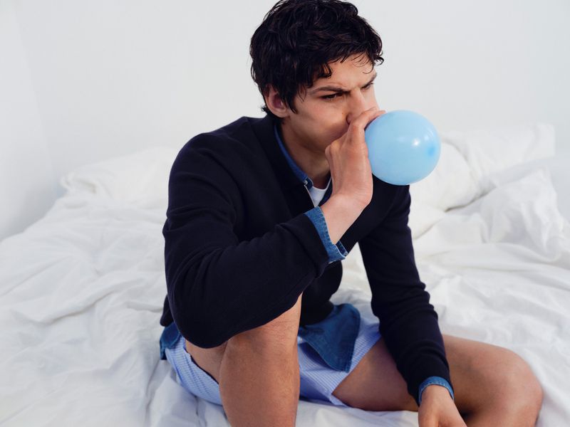 Man sitting on a white bed blowing up a light blue balloon for the Samsoe Samsoe core collection.