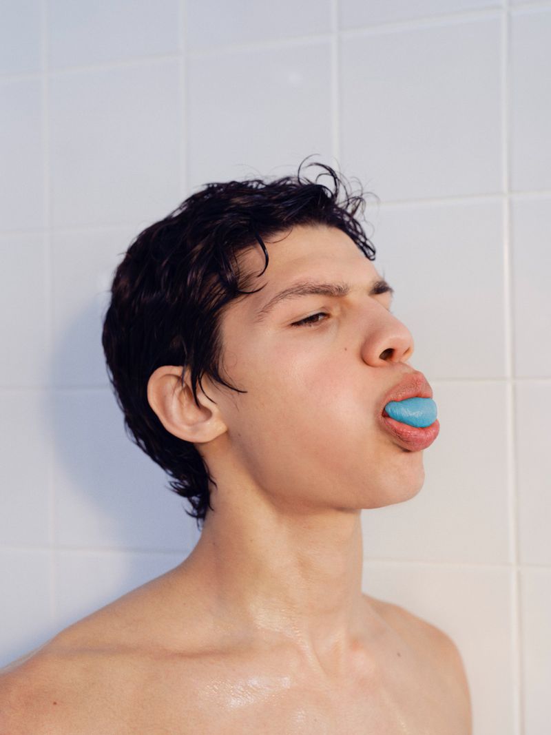 Young man with dark wet hair holding a bright blue object in his mouth against white tiled wall