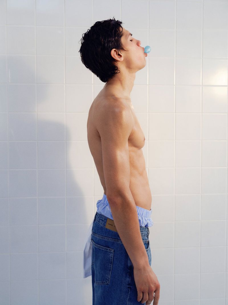 Shirtless male model in profile blowing a blue bubble gum bubble against a white tiled wall background.