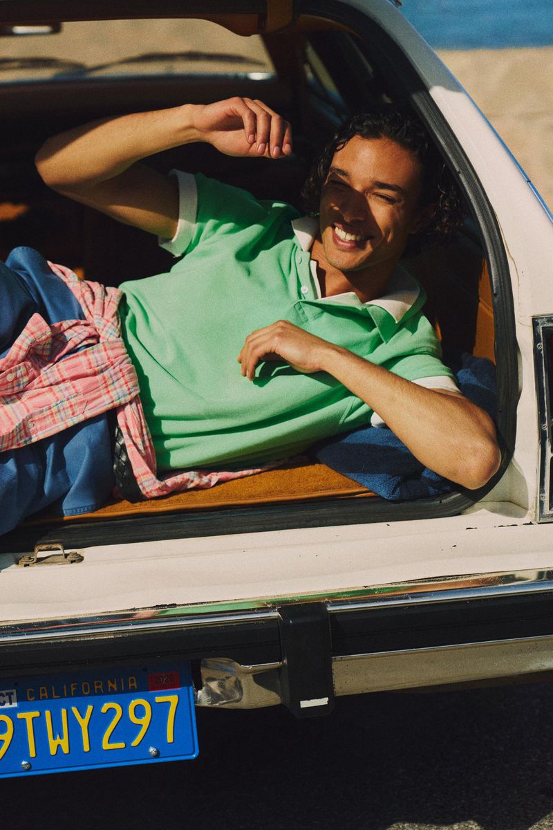 Smiling man in a green polo shirt reclining in the open trunk of a vintage car parked by the beach