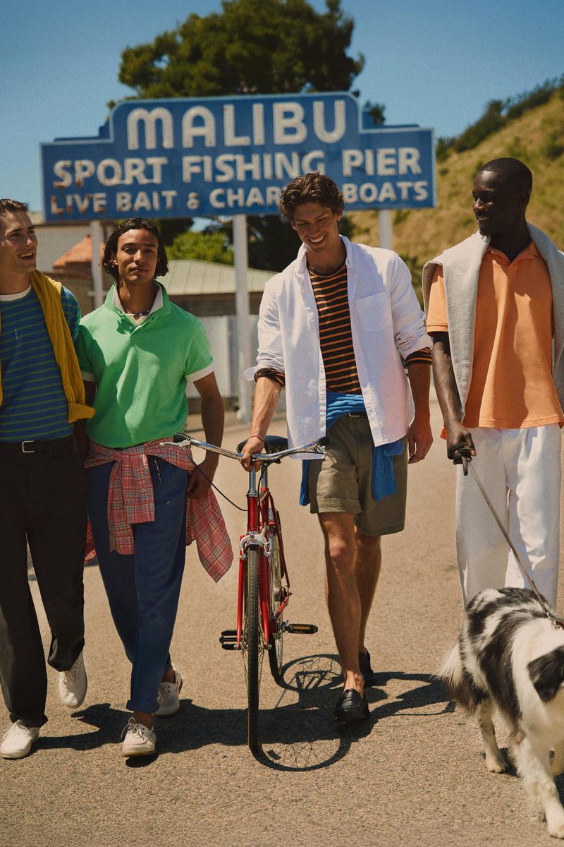 Four male models walking together outdoors near a Malibu Sport Fishing Pier sign, one pushing a red bicycle and another walking a dog.
