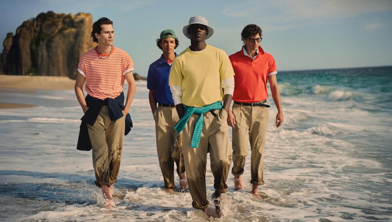 Four male models walking along the shoreline of a sandy beach wearing colorful polo shirts and trousers