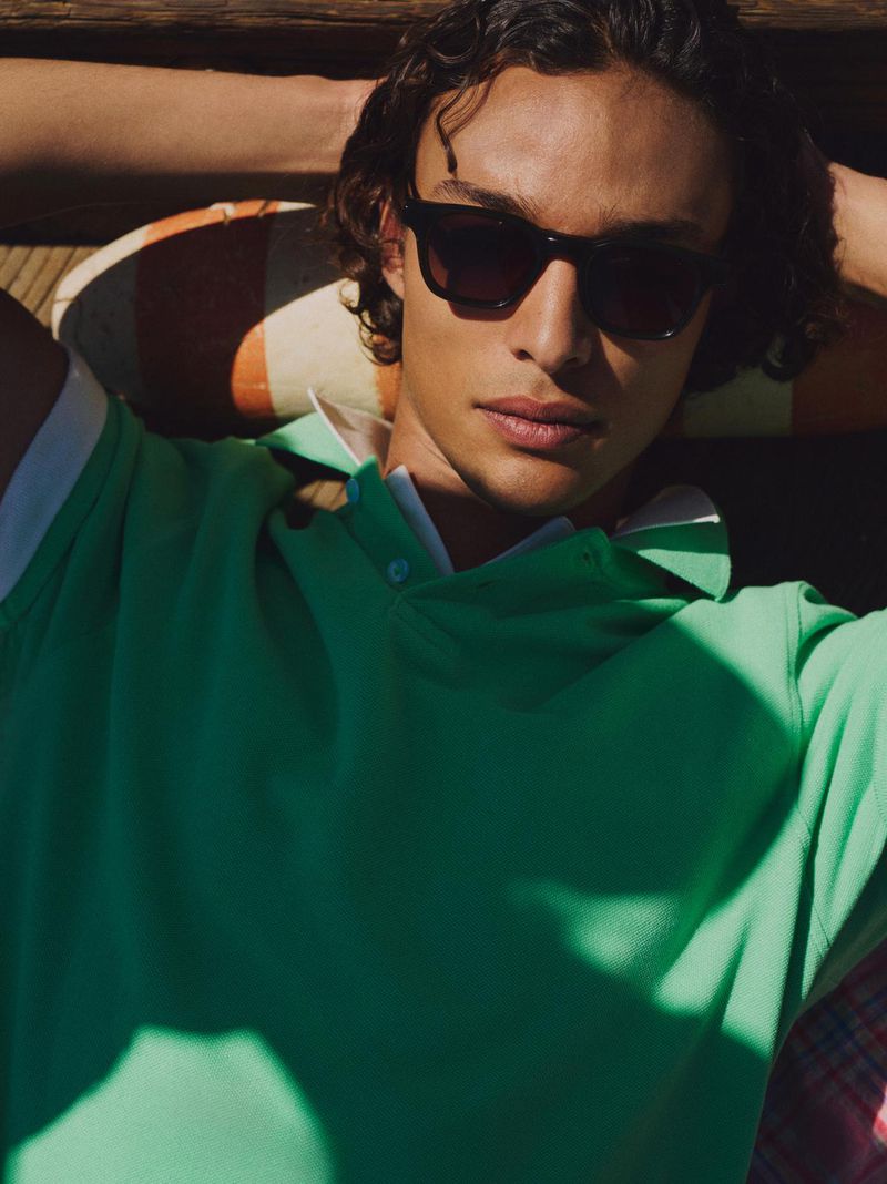 Close-up of a young adult male model wearing sunglasses and a green polo shirt, lying on a striped pillow, in an outdoor setting.