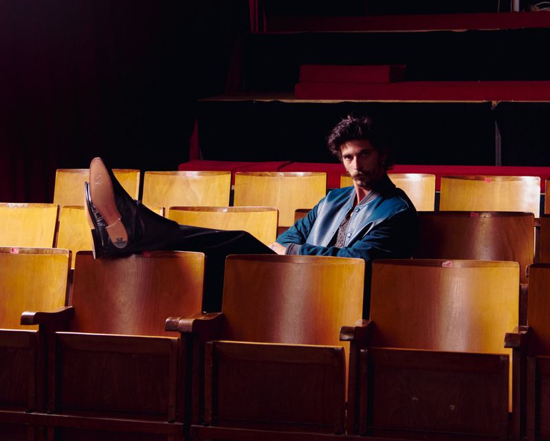 Adult male with a mustache in an empty theater wearing a dark blue jacket and black dress shoes with his feet up on the seats.