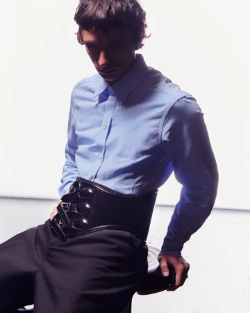 An adult male sits in a photo studio wearing a blue button-down shirt, black trousers, and a black corset-style belt.