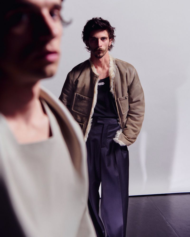 Two adult men pose indoors wearing stylish outerwear. The man in the foreground is blurred, while the other wears a suede jacket.