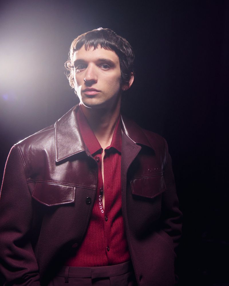 Portrait of a young adult man in a burgundy jacket and matching shirt, set against a dark backdrop with studio lighting.