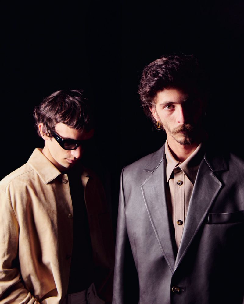 Portrait of two stylish young adult males in beige and grey blazers against a dark backdrop, one wearing dark sunglasses.