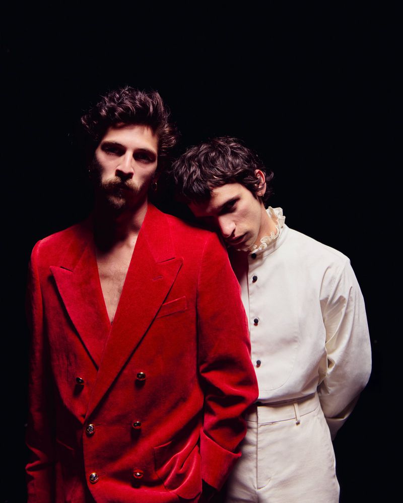 Two stylish men pose in front of a dark backdrop, one in a red velvet suit and the other in an ivory shirt with a ruffled collar.