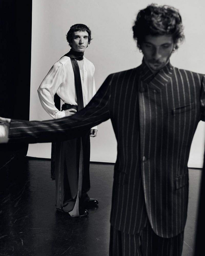Two men in pinstripe and satin outfits pose in a photo studio. Dark hair, elegant clothes. Fashion editorial shot in black and white.