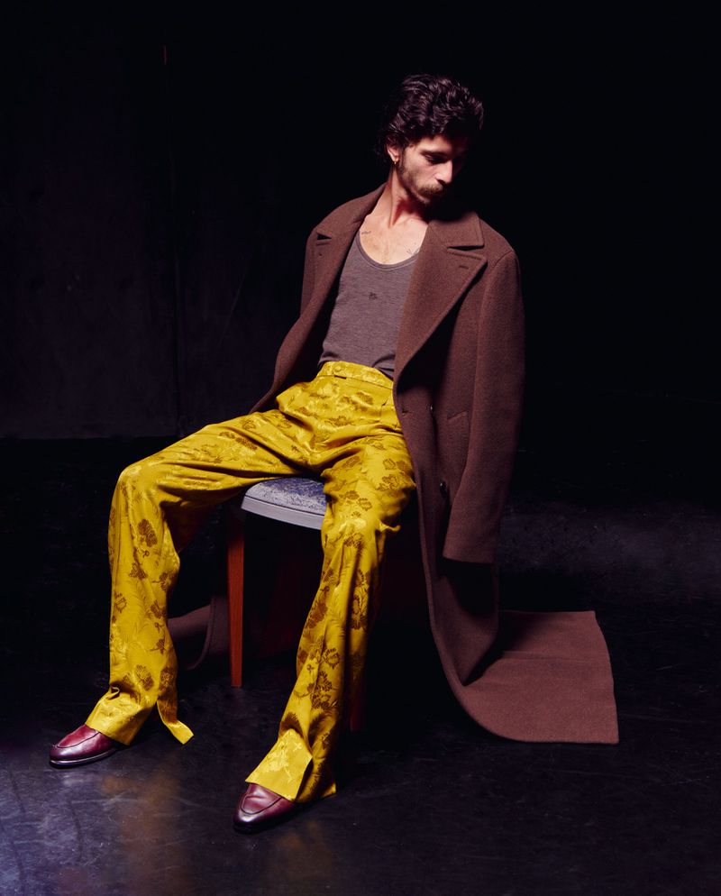 An adult male sits on a chair, wearing a brown coat, grey tank top, yellow patterned pants, and burgundy dress shoes in a dark photo studio.