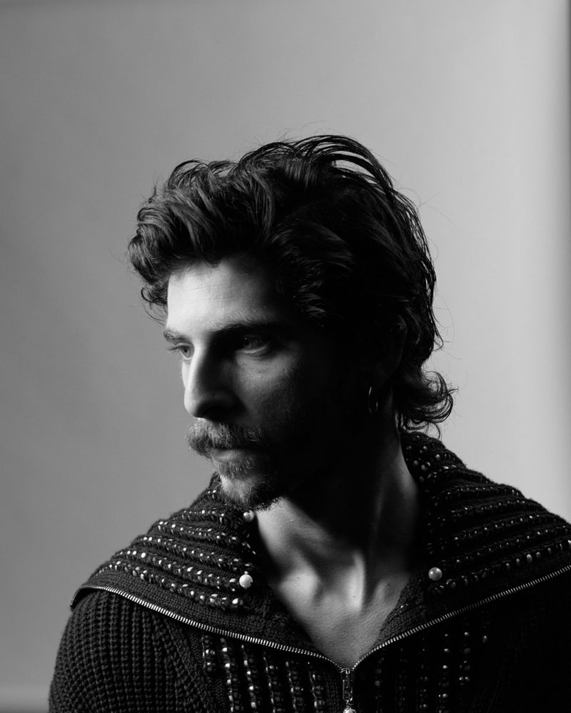 Black and white portrait of a man with a moustache wearing an embellished zippered cardigan in a studio with a neutral backdrop.