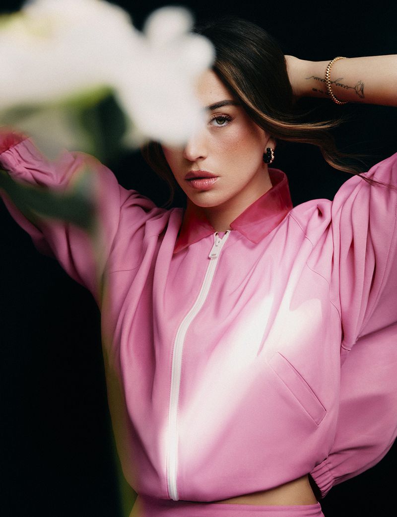 Woman wearing a pink zip jacket posing against a dark background with blurred white flowers in the foreground