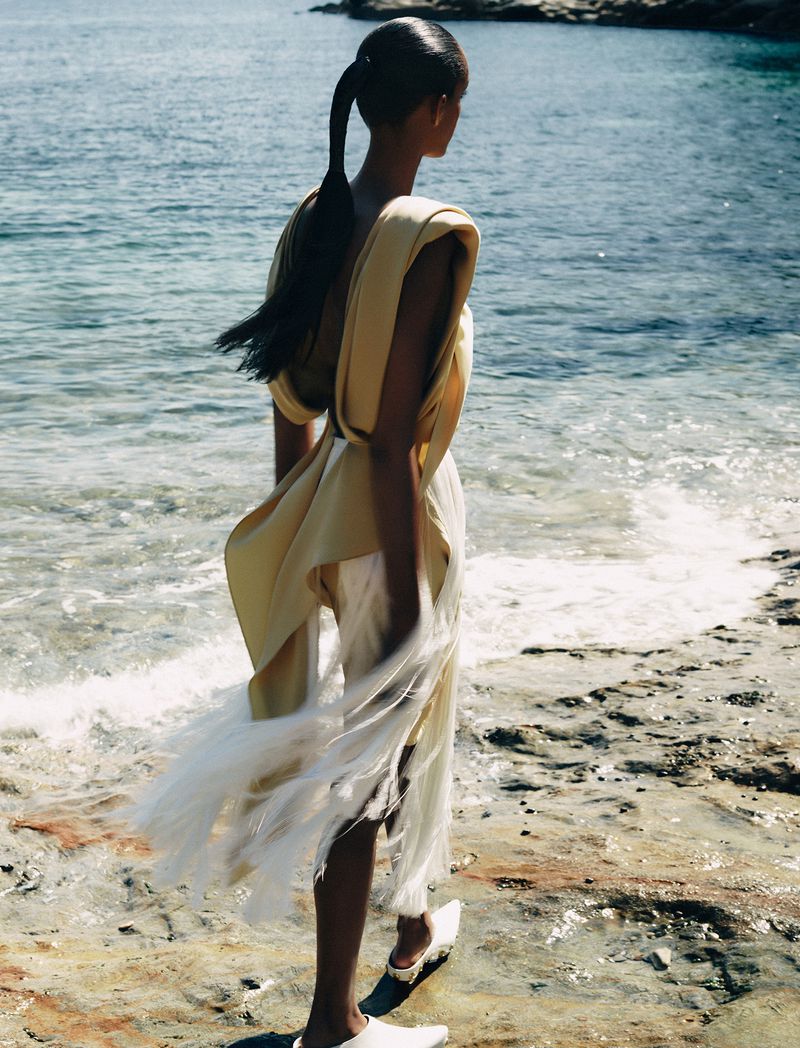Model with long dark hair in a ponytail wearing a cream draped top standing on a rocky coast near ocean water.