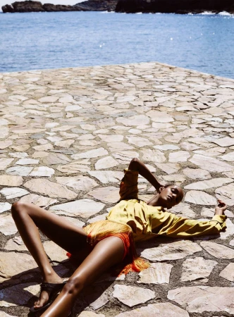 Model lying on a sunlit stone patio by the sea wearing yellow and orange fringe shorts