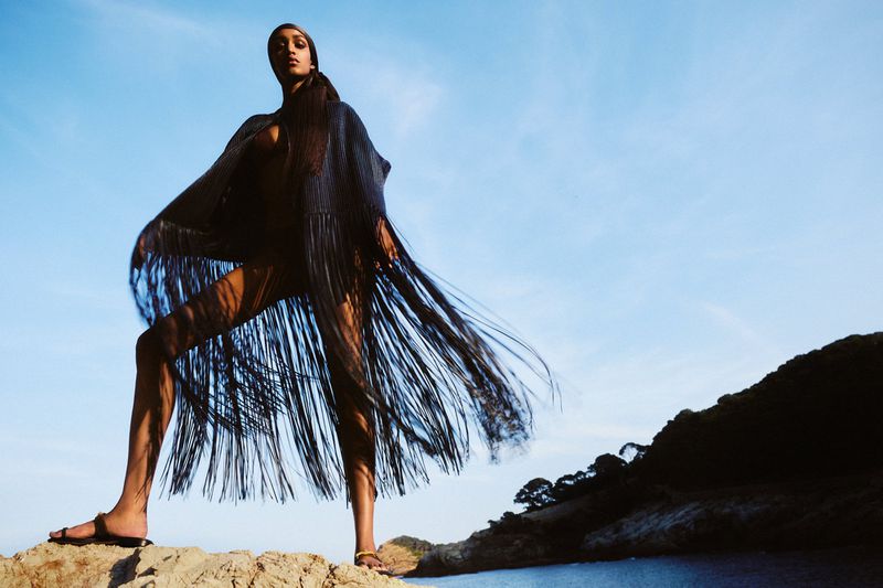 Model wearing a dark fringed cape and headscarf standing on coastal rocks against a clear blue sky