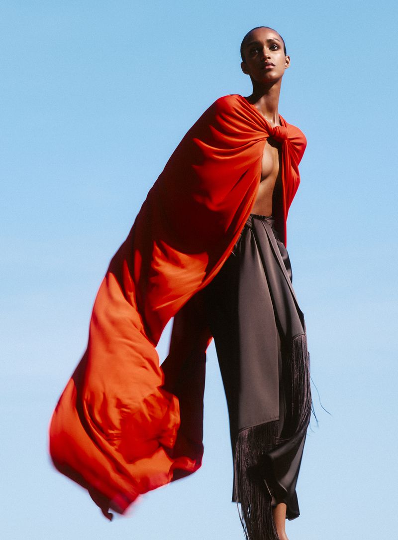 A woman in stylish black pants and a vibrant orange fabric draped over her shoulder poses against a clear blue sky, fashion forward.