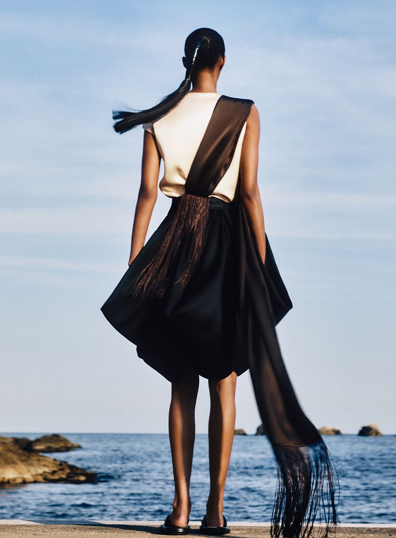 Model seen from behind standing by the ocean wearing a cream top and dark skirt with a long flowing sash.
