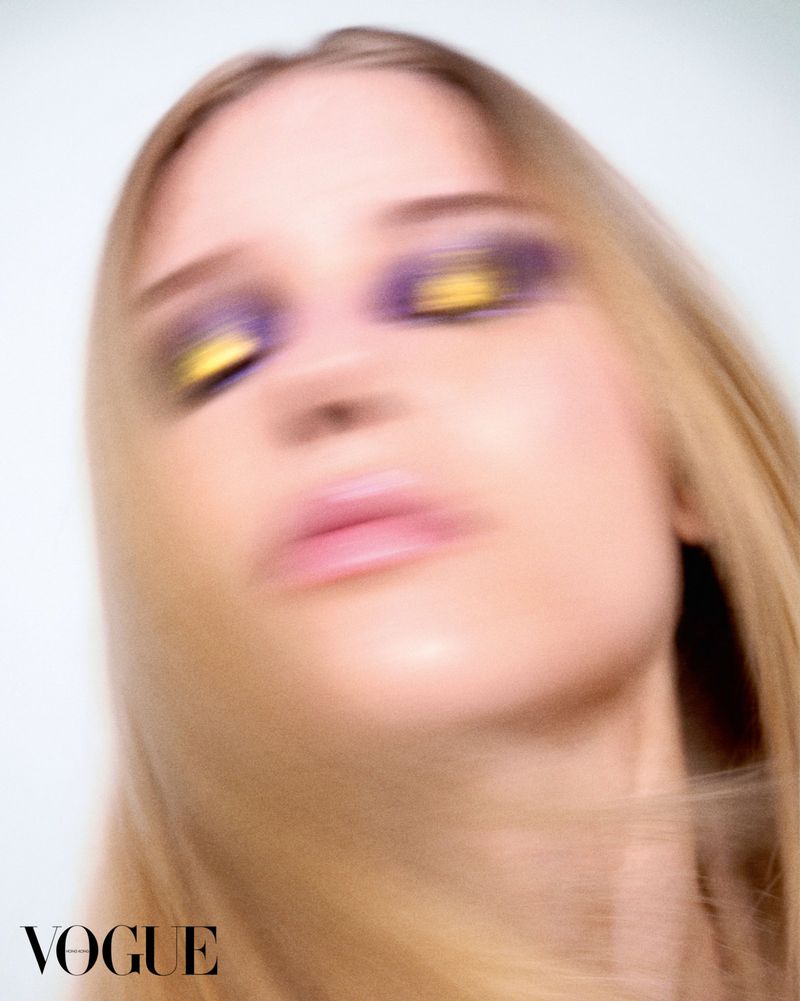 Close-up portrait of a blonde model with motion blur, wearing purple and gold eyeshadow and pink lipstick against a white background.