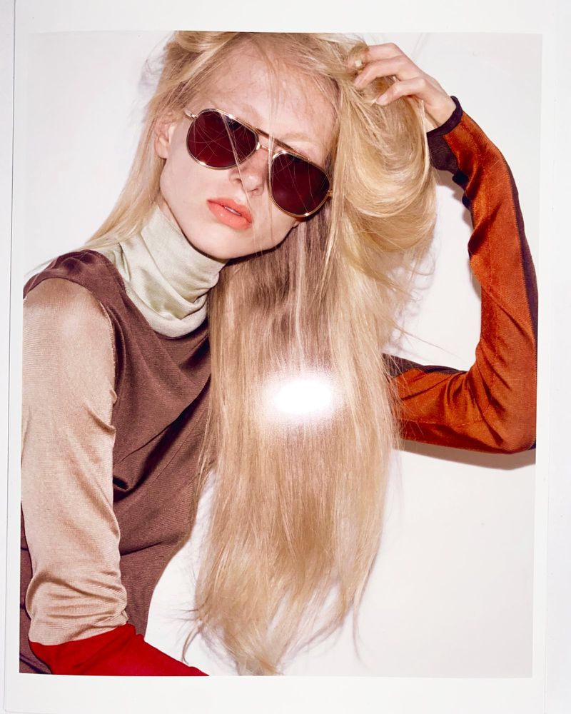 Blonde model wearing aviator sunglasses and brown turtleneck top against white backdrop