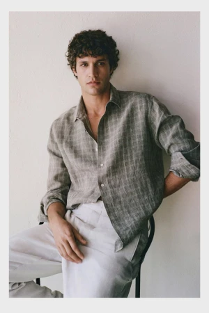 Male model Francisco Henriques wearing a grey patterned linen shirt and light trousers seated against a textured white wall.